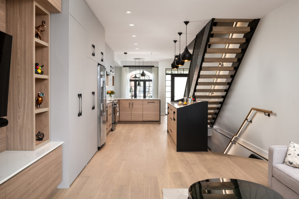 Modern kitchen and living space with light wood floors, built-in shelves, sleek cabinets, black pendant lights, and a floating staircase with glass railing, leading to a bright area with large windows.