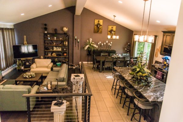Modern open-concept living space with a neutral-toned living room, a long dining table, and a kitchen with a granite island and barstools. Decor includes wall art, plants, and shelves with various items.