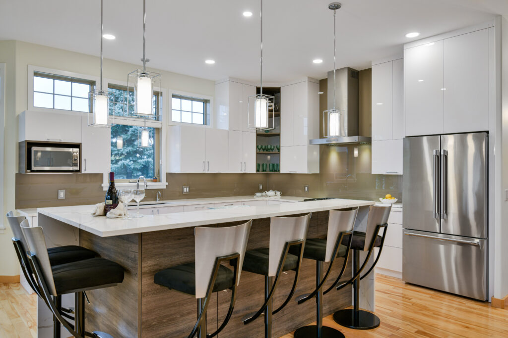 Modern kitchen with a large island, four barstools, pendant lights, stainless steel appliances, white cabinets, and wooden floors. There is a bottle of wine and glasses on the island, and a window view in the background.