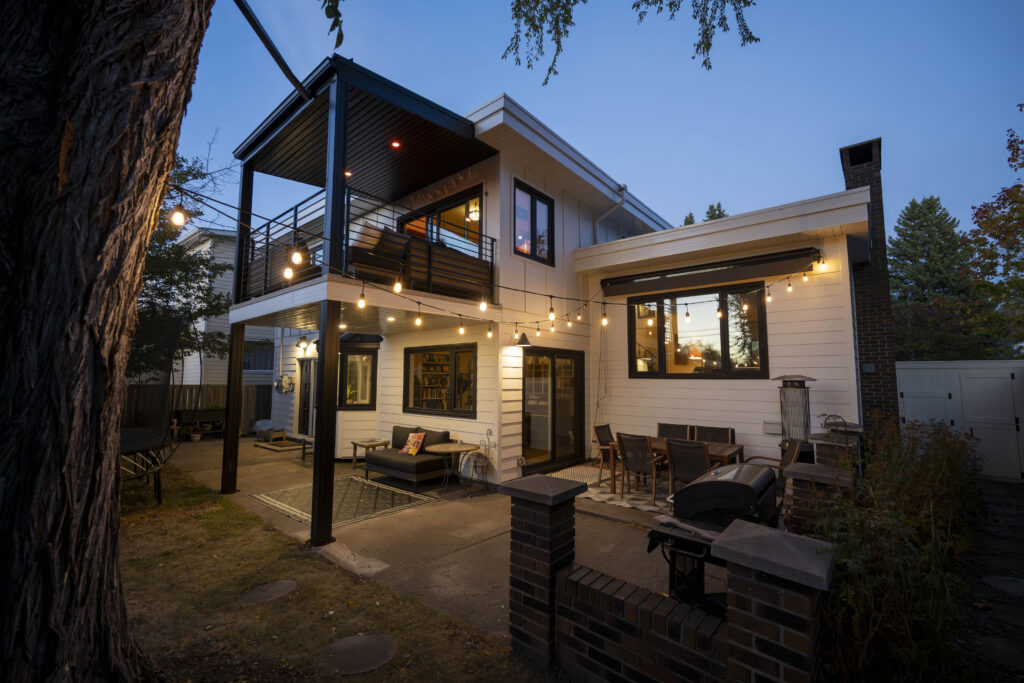 Modern two-story house at dusk with outdoor seating areas, string lights, a balcony, large windows, and a brick patio, surrounded by trees and a fenced yard.