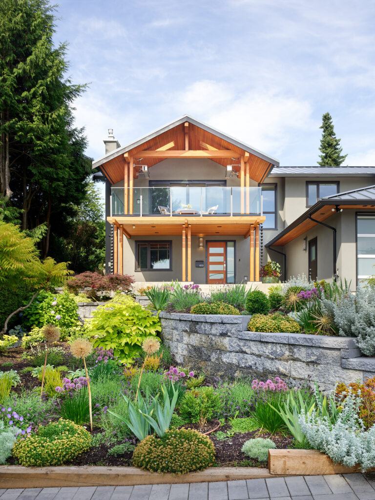 A modern two-story house with large windows, a glass balcony, and wooden accents, surrounded by a lush, landscaped garden with colorful flowers, shrubs, and stone retaining walls.