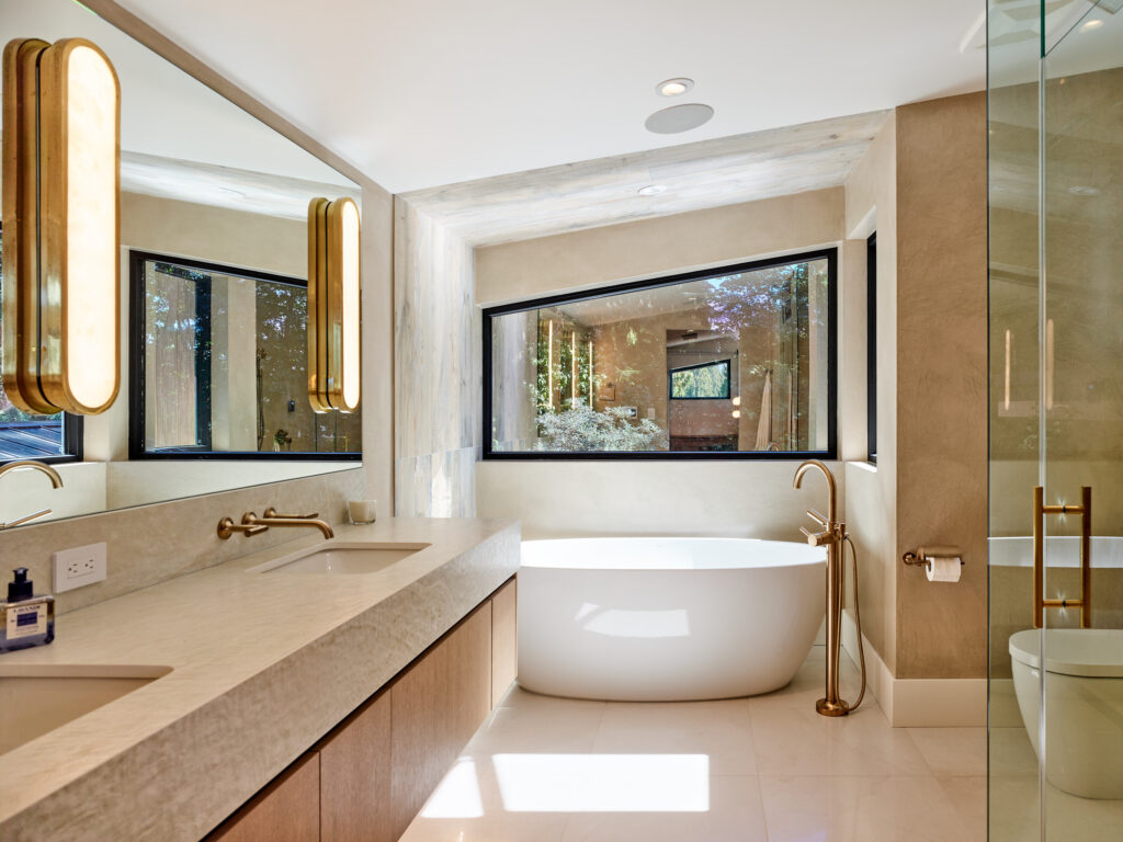 Modern bathroom with large mirror, double sink vanity, freestanding white bathtub by a window, gold fixtures, beige walls, glass shower enclosure, and soft overhead lighting.
