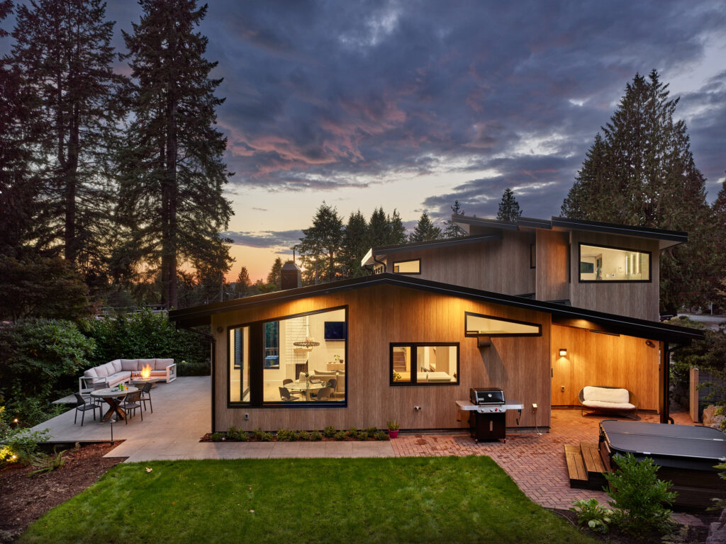 A modern two-story house with large windows and wooden siding, surrounded by tall trees. The backyard features a patio with seating, a barbecue grill, a hot tub, and a well-kept lawn at sunset.