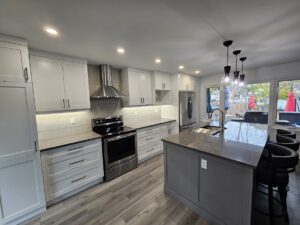 Modern kitchen with light gray cabinets, stainless steel appliances, a center island with black barstools, pendant lights, and large windows overlooking a backyard with red patio umbrellas.