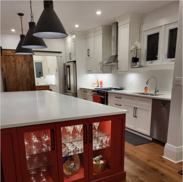 Modern kitchen with white cabinets, stainless steel appliances, a large island with a red base featuring glass doors displaying glassware, and pendant lights hanging above. Wooden floor and sliding barn door are visible.