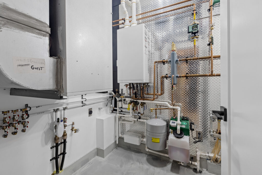A clean utility room with exposed copper pipes, a water heater, control boxes, a filtration system, and various plumbing fixtures mounted on white walls and a metallic panel. The concrete floor and organized layout are visible.