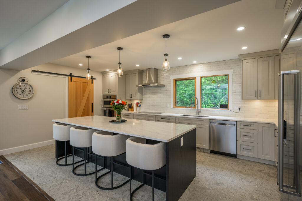 Modern kitchen with a large island, four cushioned stools, pendant lights, stainless steel appliances, light gray cabinets, a farmhouse sink under a window, and a sliding barn door next to a wall clock.