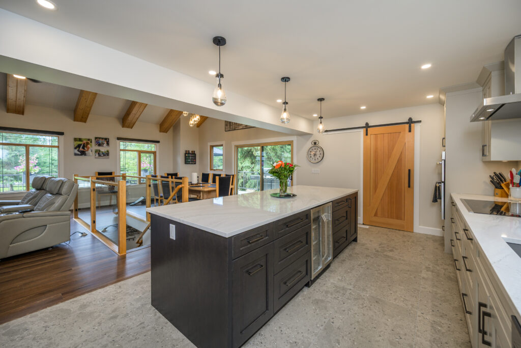 Modern kitchen with a large dark island, white countertops, pendant lights, and a wooden sliding barn door. Dining and living areas with large windows and exposed beams are visible in the background.