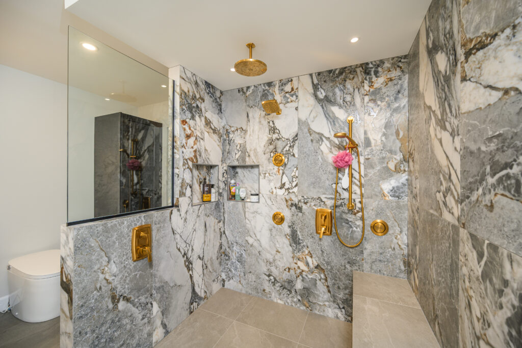 A modern bathroom features a spacious marble shower with gold fixtures, a glass partition, built-in shelves with toiletries, and a bench; a toilet is visible in the background.