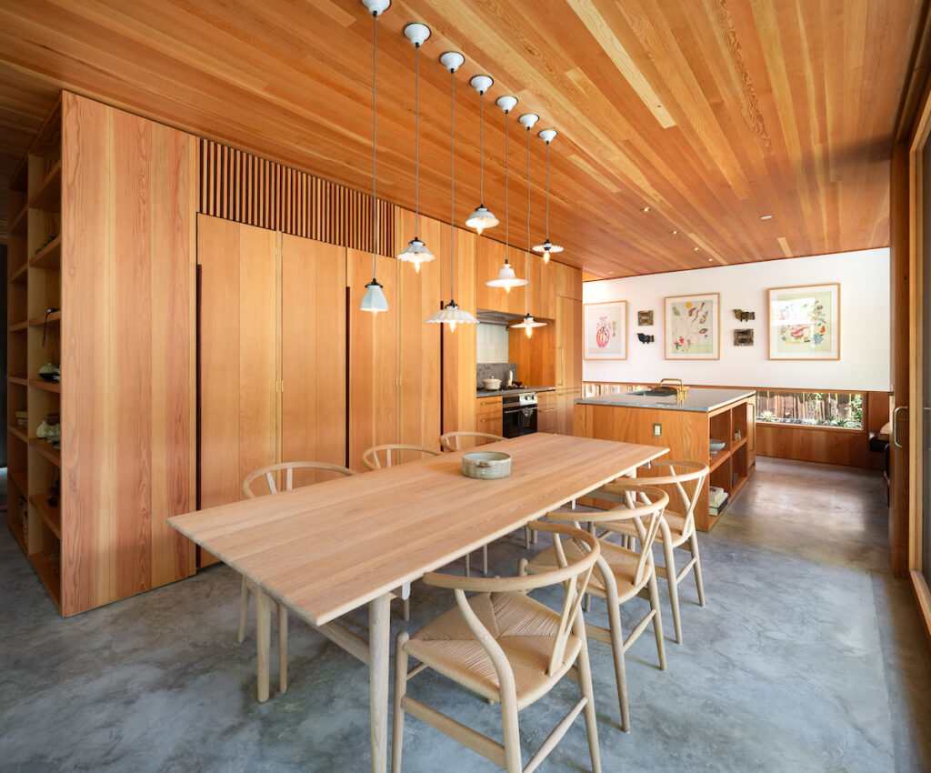 A modern kitchen and dining area with wooden walls, ceiling, and furniture. Pendant lights hang above a large dining table with six chairs. The kitchen has open shelving, framed art on the wall, and a polished concrete floor.