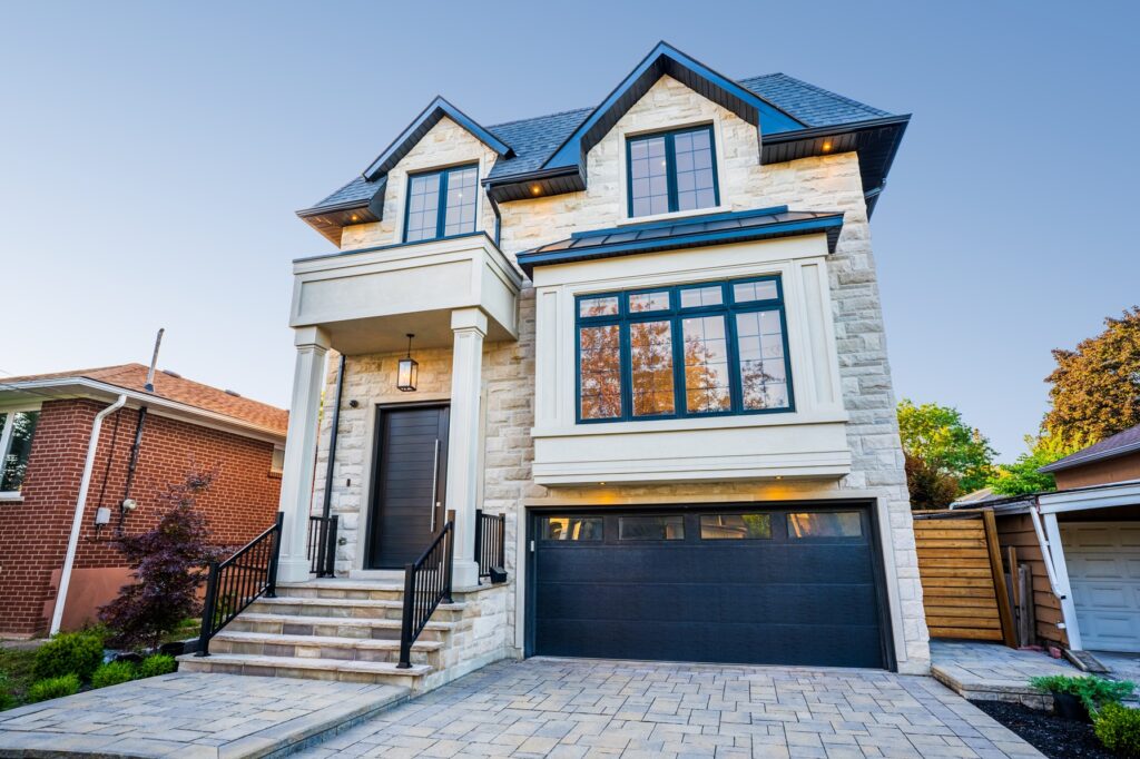 A modern, two-story stone house with large black-framed windows, a black double garage door, front steps with railings, and a paved driveway, surrounded by other houses and trees.