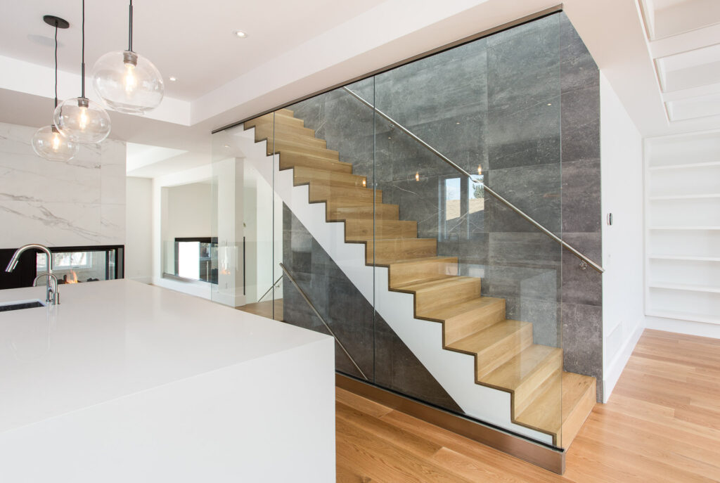 A modern staircase with wooden steps and glass railings is set against a gray stone wall in a bright, minimalist interior with white walls, hanging glass pendant lights, and light wood flooring.