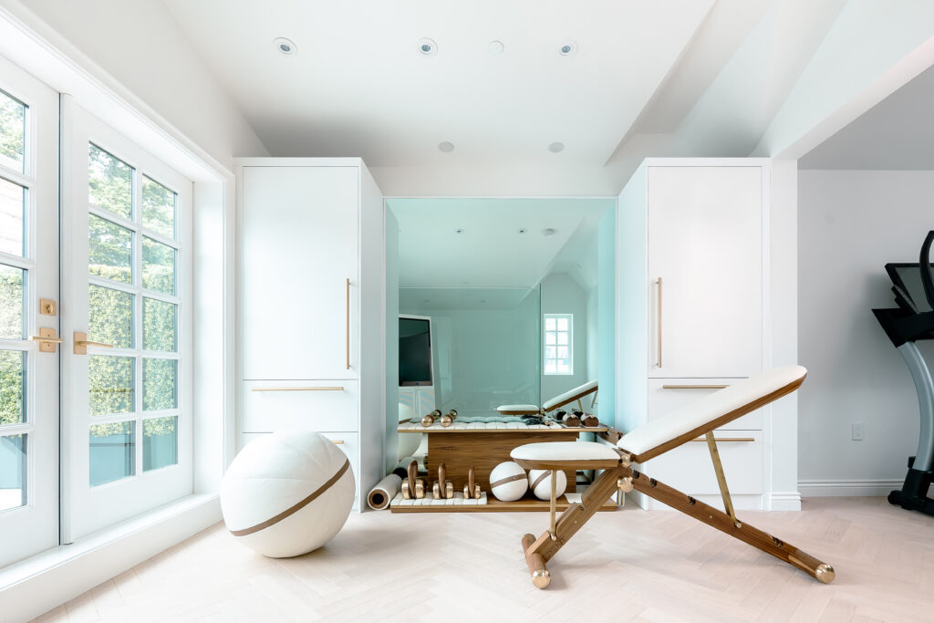 A bright home gym with large windows, white cabinets, a mirror wall, an exercise ball, wooden gym equipment, and an adjustable white workout bench on light wood flooring.