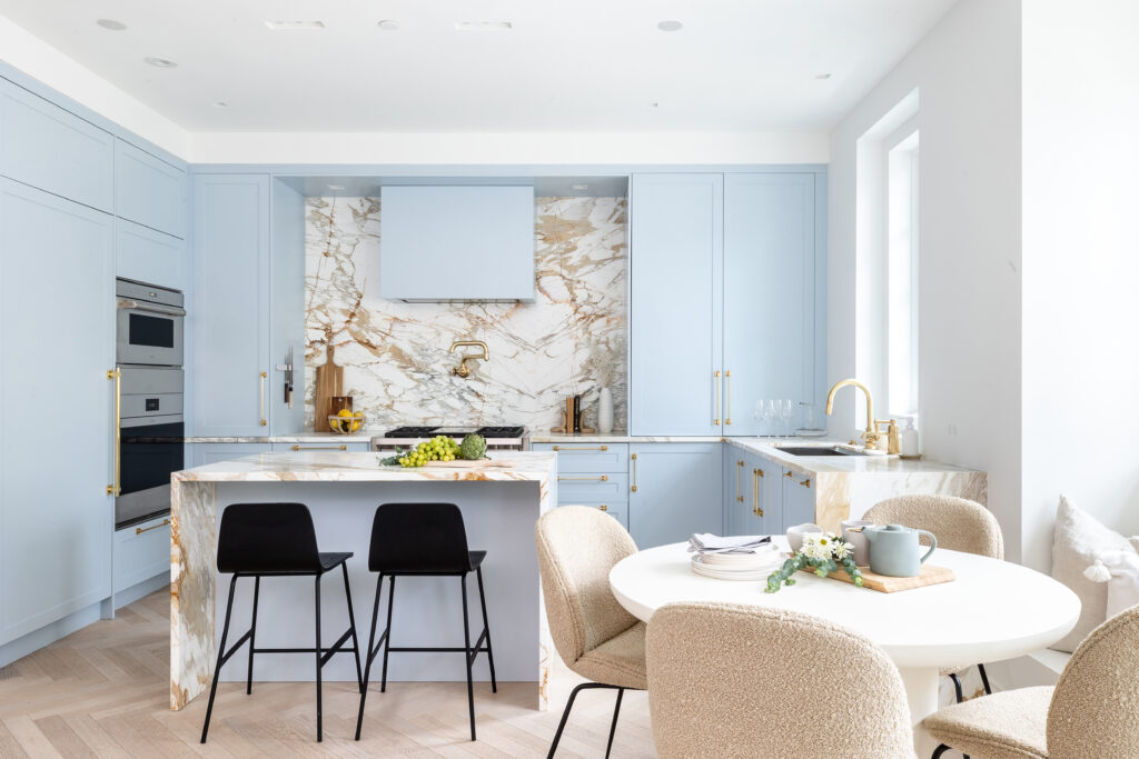 Modern kitchen with light blue cabinets, marble backsplash and island, built-in stainless steel appliances, black and beige chairs, round dining table with place settings, and bright natural lighting.
