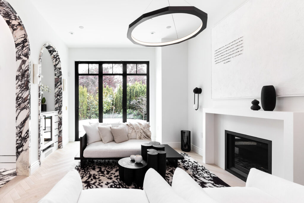 A modern, minimalist living room with white walls, black accents, large windows, a black-and-white marble archway, abstract art, a contemporary fireplace, and white furniture on a patterned rug.