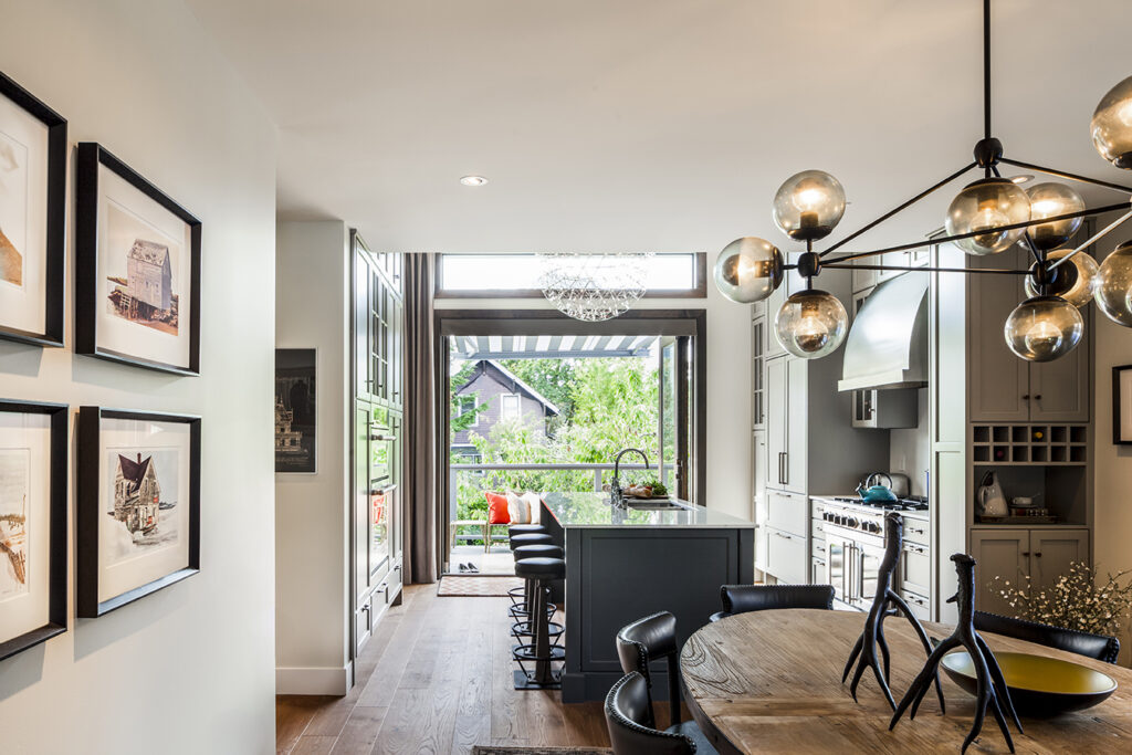 A modern kitchen and dining area with large windows, stylish lighting fixtures, a round dining table, black chairs, and an island with stools. Outdoor greenery is visible through the open glass doors.