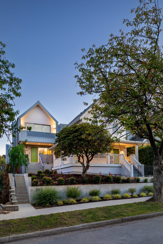 A modern two-story house with large windows, a green front door, and a spacious porch is surrounded by trees and landscaped greenery, viewed from the sidewalk at dusk.