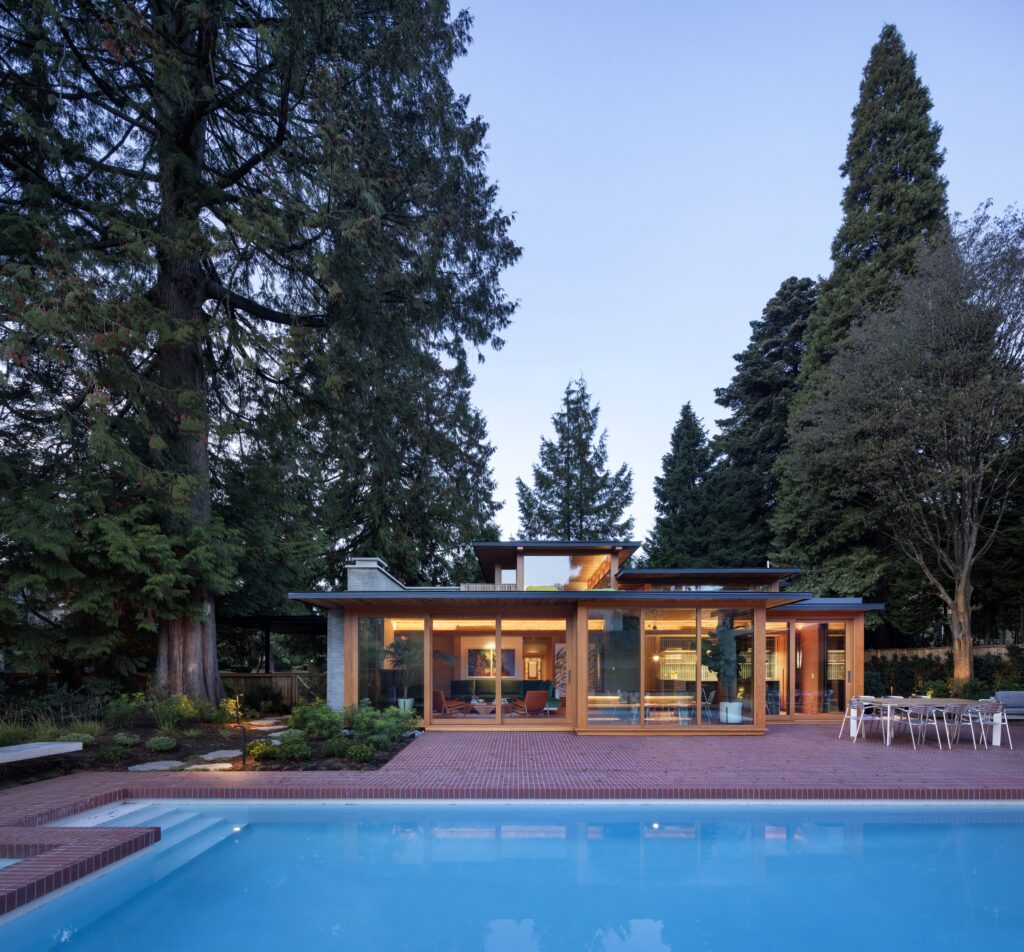 A modern house with floor-to-ceiling windows and warm interior lights stands behind a large, clear swimming pool, surrounded by tall trees and a brick patio at dusk.