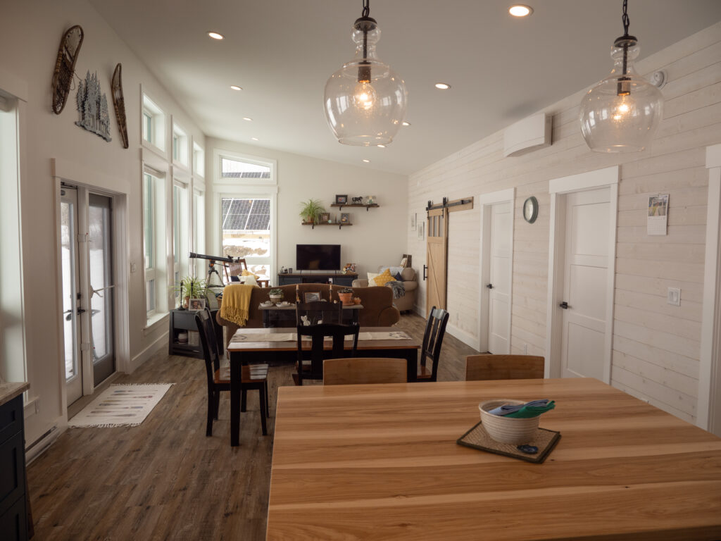 A modern open-concept living and dining area with wooden floors, large windows, a wooden dining table, cozy sofa, TV, and sliding barn door. Pendant lights hang above a wooden kitchen island in the foreground.