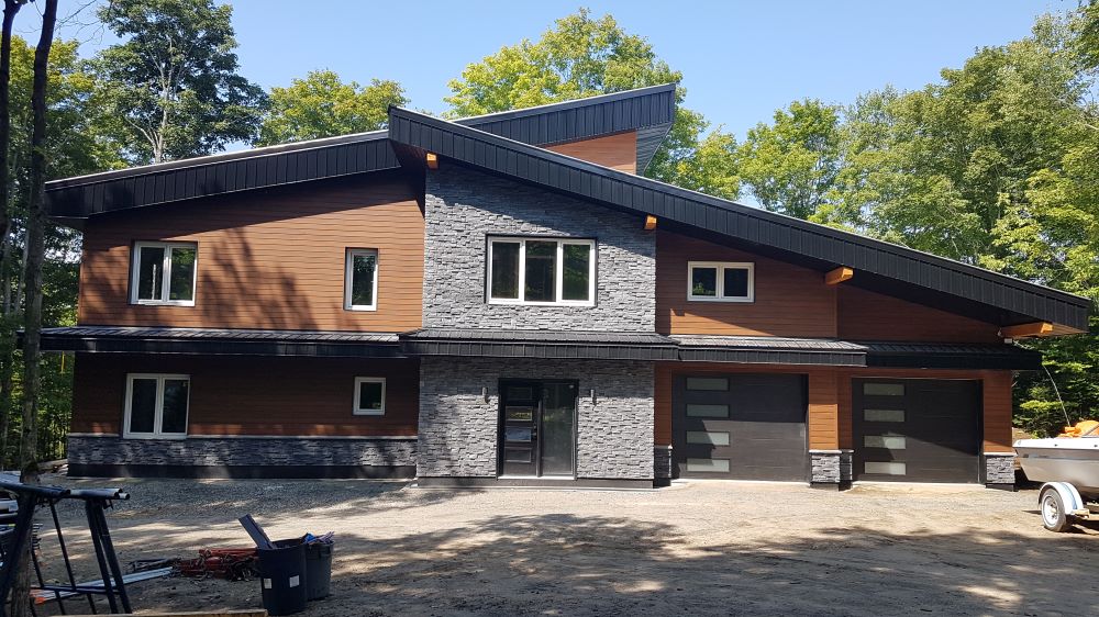 A modern two-story house with wood and stone siding, large windows, and a sloped roof, featuring two garage doors. The house is surrounded by tall green trees under a clear blue sky.