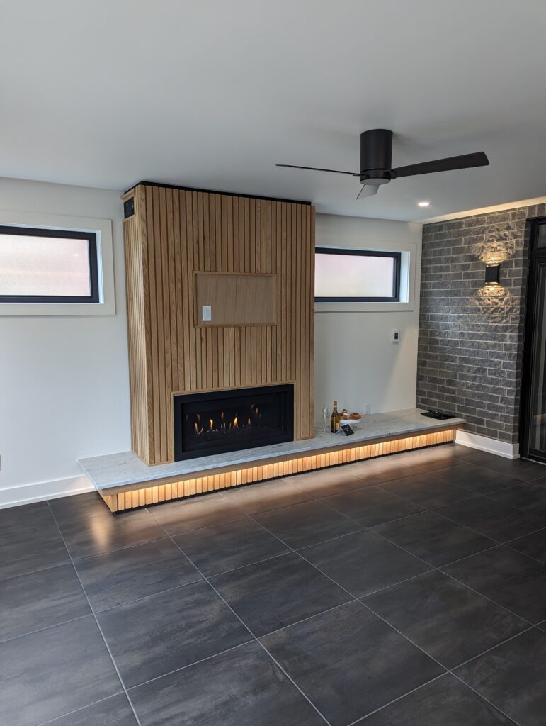 Modern living room with a black tile floor, a wood-paneled fireplace with a built-in gas fire, LED strip lighting under a stone hearth, two horizontal windows, and a ceiling fan with black blades.