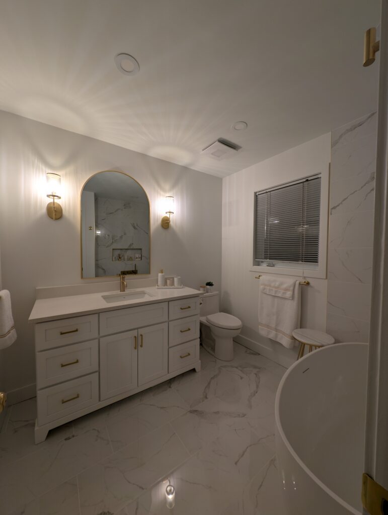 A modern bathroom with white marble floors, a white vanity with gold accents, a large arched mirror, two wall sconces, a toilet, a towel rack, and a freestanding bathtub.