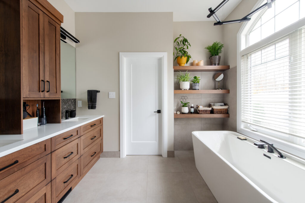 Modern bathroom with wooden cabinets, a white bathtub, floating shelves with plants and decor, large window with blinds, and a white door centered between the vanity and the tub. The space is bright and neatly organized.