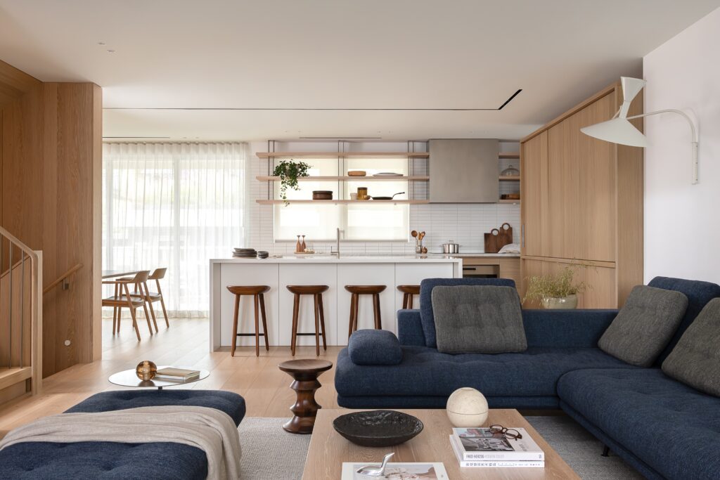 A modern open-plan living room and kitchen with wooden floors and cabinetry, navy blue sofas, a wooden coffee table, bar stools at a kitchen island, and large windows with sheer curtains letting in natural light.