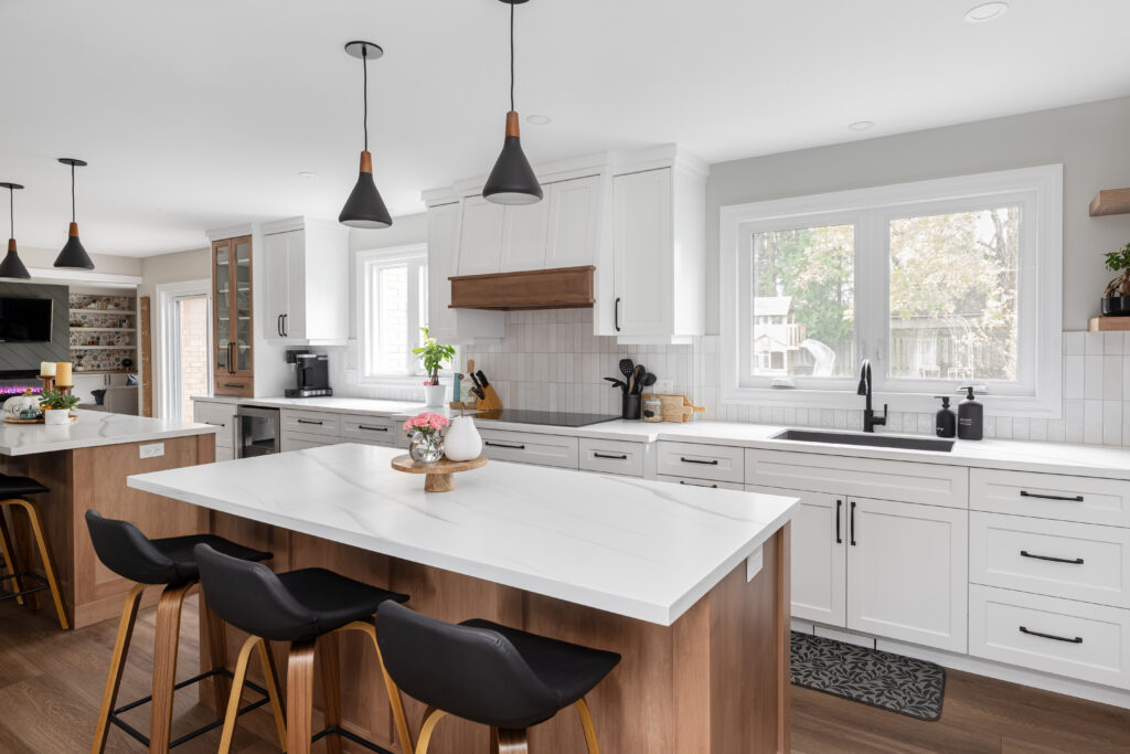 Modern kitchen with white cabinets, wood accents, a large island with barstools, black pendant lights, and large windows letting in natural light. The counters are clean and decorated with a few kitchen items.