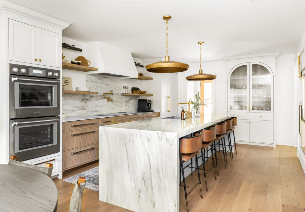 Modern kitchen with a large marble island, gold pendant lights, brown leather bar stools, wood flooring, double ovens, marble backsplash, and white cabinetry with brass accents.