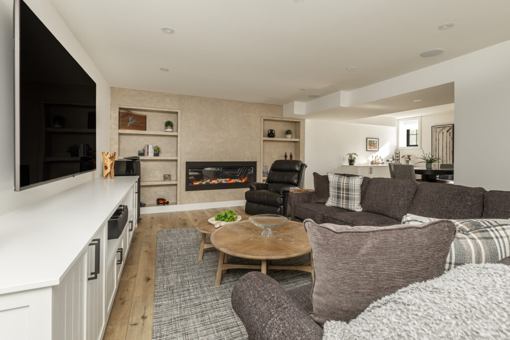 A modern living room with a large wall-mounted TV, dark brown sectional sofa, round wooden coffee table, electric fireplace, built-in shelves, and light wood flooring. A dining area is visible in the background.