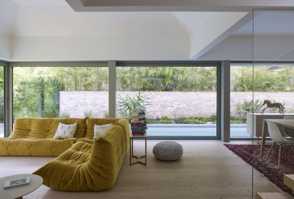 A modern living room with a large yellow sofa, a small stack of books on a side table, a round gray pouf, and floor-to-ceiling windows overlooking a garden with a pool and brick wall.