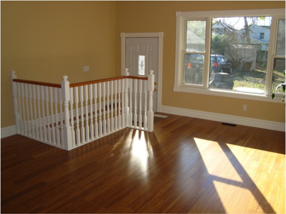 A sunlit, empty room with light wood floors, beige walls, a white railing around a stairway, a white door, and a large window showing a view of a car and trees outside.