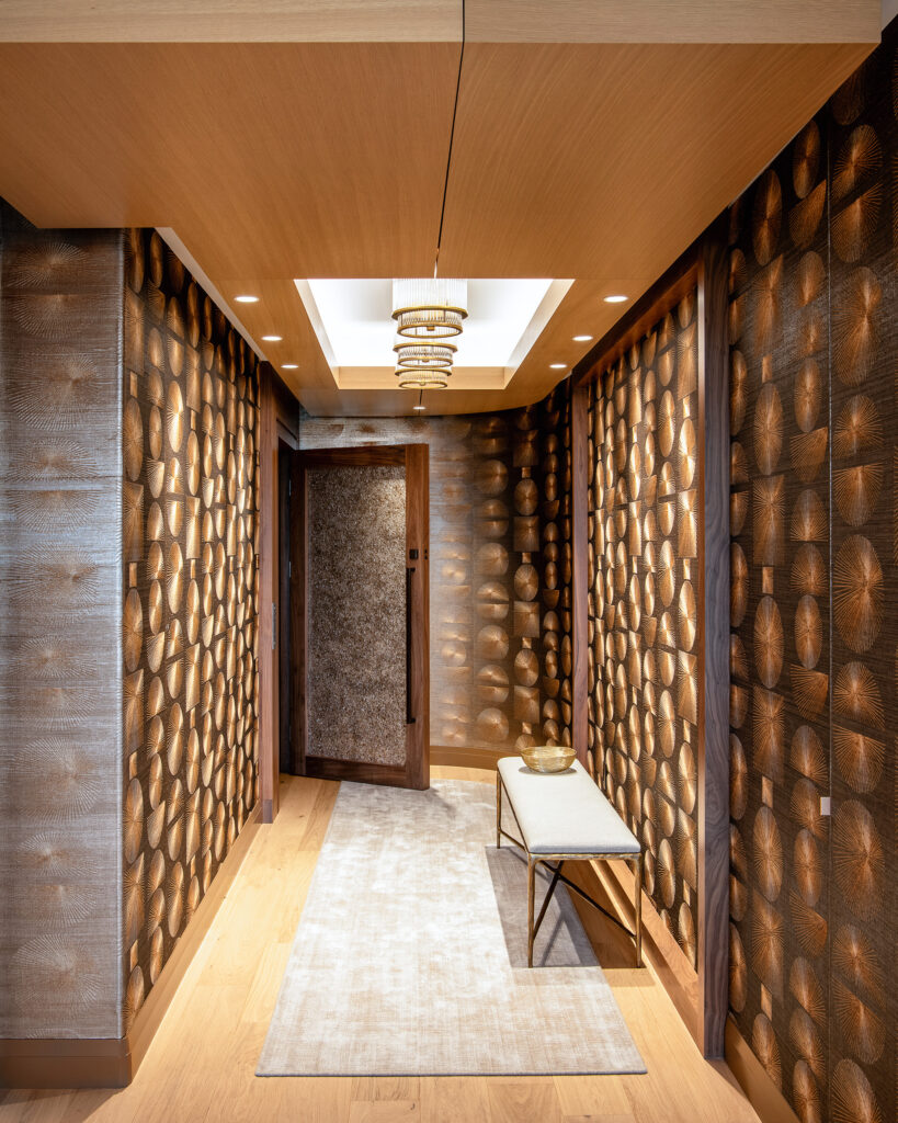 A modern hallway with patterned brown walls, a light wood floor, a beige rug, a cushioned bench with a gold bowl, and a geometric ceiling light fixture under a recessed ceiling.