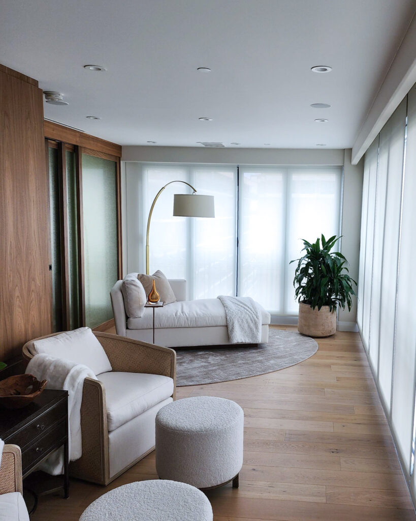 Modern living room with light wood floors, white lounge chairs, a cushioned chaise, round ottomans, floor lamp, large potted plant, and sheer curtains letting in natural light.