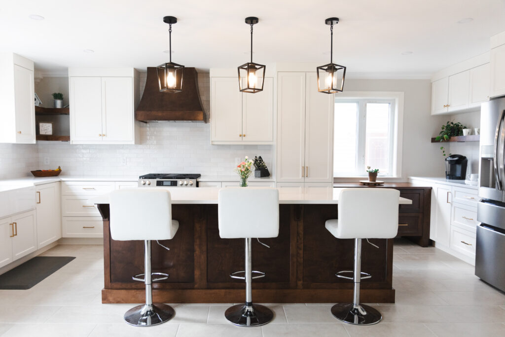 Modern kitchen with white cabinets, a dark wood island, three white barstools, pendant lights, stainless steel appliances, and a vase with flowers on the island. Bright and clean with natural light from a window.
