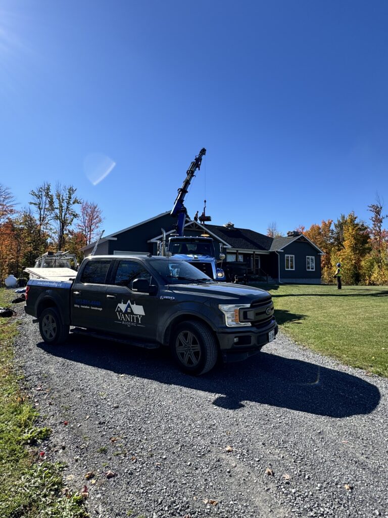 A black pickup truck with Vanity Roofing logo is parked on a gravel driveway in front of a dark-colored house, where workers and a crane are on the roof under a clear blue sky and surrounded by autumn trees.