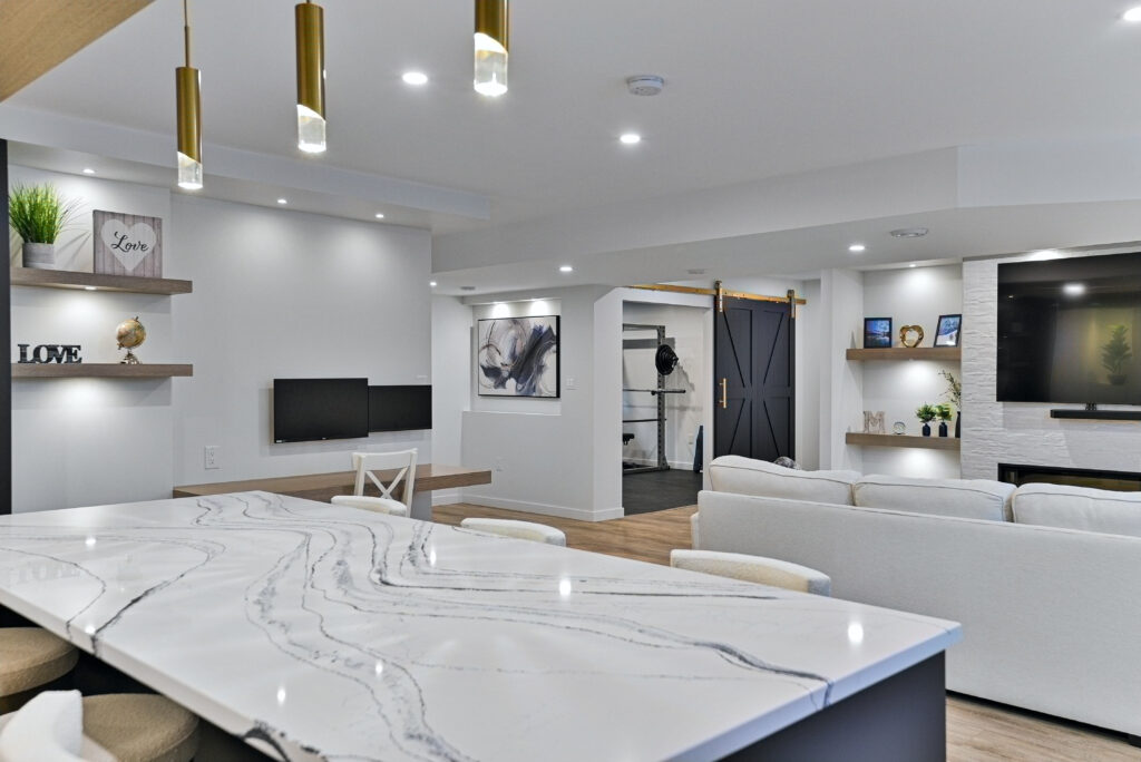 Modern open-concept living space with white walls, marble kitchen island, pendant lights, cozy sofa, wall-mounted TVs, floating shelves with decor, and a sliding barn door leading to another room.