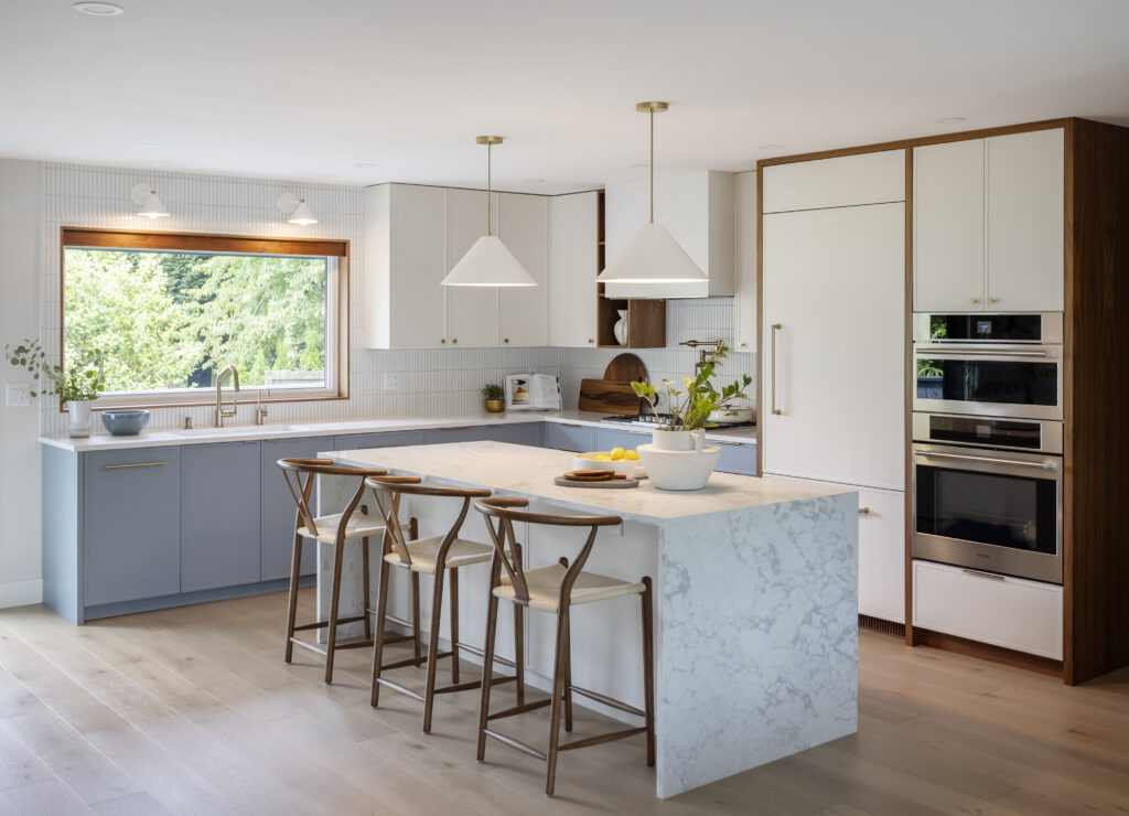 Modern kitchen with large marble island, four wooden bar stools, white and light blue cabinets, stainless steel appliances, pendant lights, and a large window overlooking greenery. A bowl of lemons sits on the island.