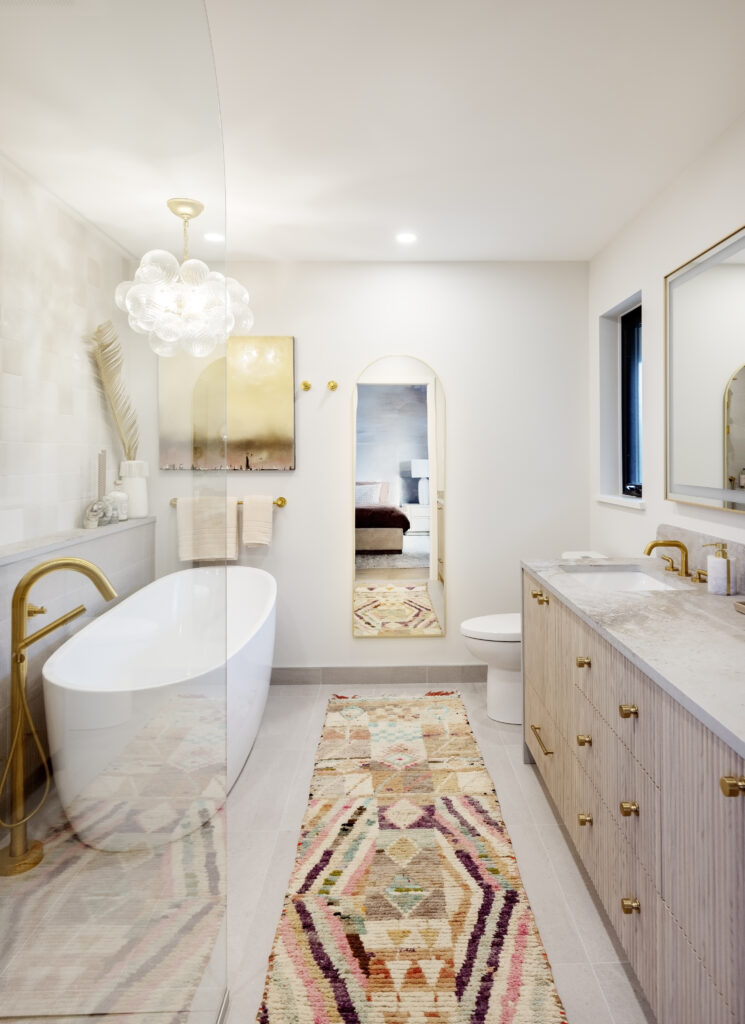 Modern bathroom with a freestanding white tub, gold fixtures, a double vanity, patterned rug, arched mirror, abstract wall art, and a glass shower. The space is bright with neutral tones and elegant decor.