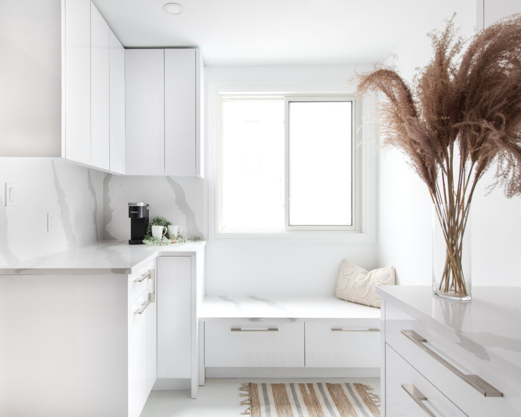 A bright, modern kitchen with white cabinets, marble countertops, a window seat with cushions, a striped rug, and a tall vase filled with dried pampas grass on a white dresser.
