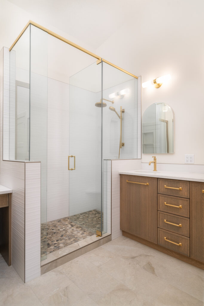 Modern bathroom with a glass-enclosed shower featuring gold fixtures and a pebble tile floor, next to a wooden vanity with gold handles and a matching faucet under a wall-mounted light and mirror.