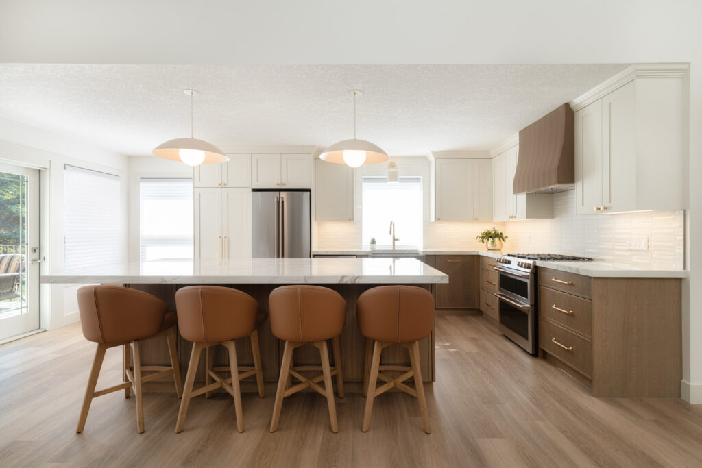 A modern kitchen with light wood floors, white cabinets, a large island with marble countertop, five brown barstools, stainless steel appliances, and pendant lights overhead. Natural light fills the space.
