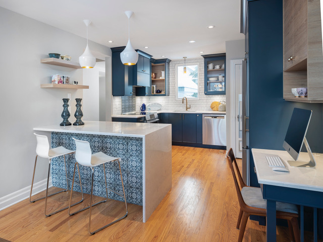 Modern kitchen with blue cabinets, white countertops, and wood flooring. An island with patterned tiles has two white barstools. Pendant lights hang above. A built-in desk with a computer is visible on the right side.