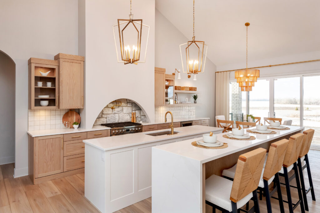Bright, modern kitchen featuring a large white island with barstools, gold pendant lights, light wood cabinets, a built-in oven with stone backsplash, open shelving, and a dining area with large windows overlooking an outdoor view.