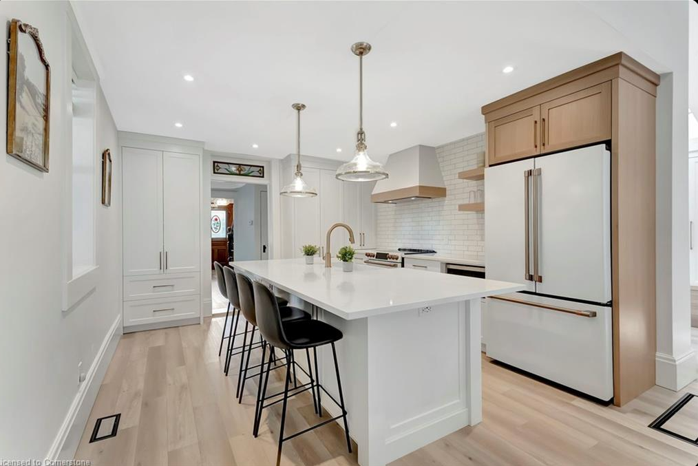 Modern kitchen with a large white island, four black bar stools, pendant lights, light wood flooring, white cabinetry, and a refrigerator with wood accents. The space is bright and minimalist.