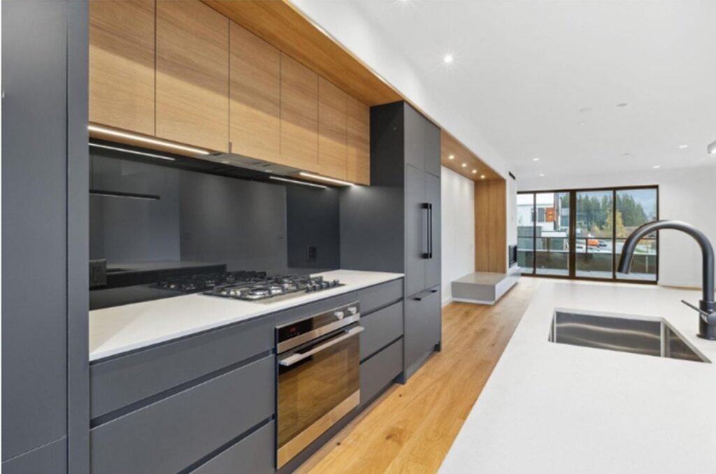 Modern kitchen with sleek black and wood cabinets, built-in appliances, a gas stove, and a large island with a sink. Floor-to-ceiling windows provide natural light and a view of trees outside.