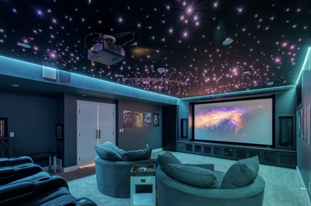 A modern home theater room with plush seating, a large screen displaying a galaxy scene, and a ceiling decorated with glowing star-like lights and a ceiling projector. Blue LED lighting lines the upper walls.