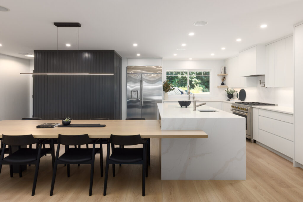 Modern kitchen with white cabinets, a marble island with integrated sink, stainless steel appliances, light wood flooring, a long wooden dining table with six black chairs, and large window letting in natural light.