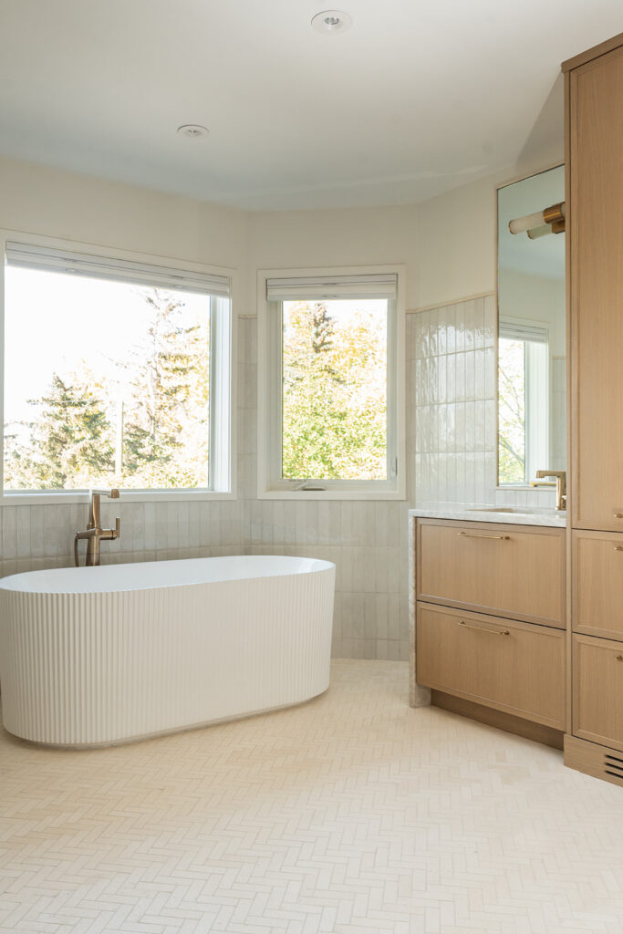 A modern bathroom with a white freestanding bathtub, large windows with views of trees, light wood cabinetry, a marble countertop, and neutral tile flooring. The space is bright and minimalistic.